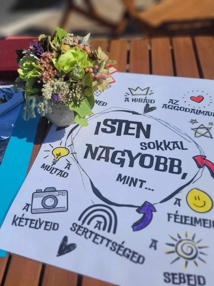Kézműves asztal mezei virágcsokorral és az „Isten sokkal nagyobb, mint…” feliratú tablóval, mellette hegyi táj képeslapja és Biblia